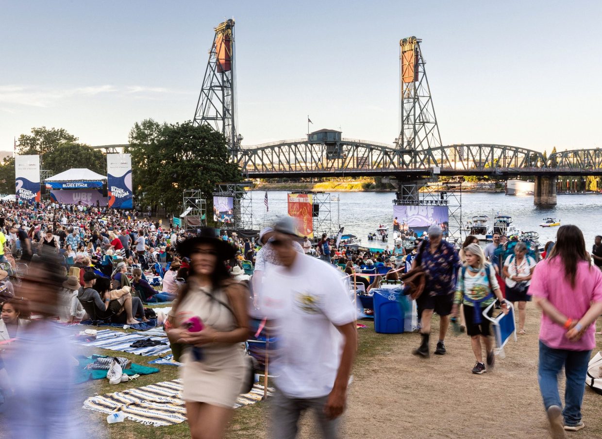 Crowds gather at the Waterfront Blues Festival beside the river, with a raised steel bridge in the background.