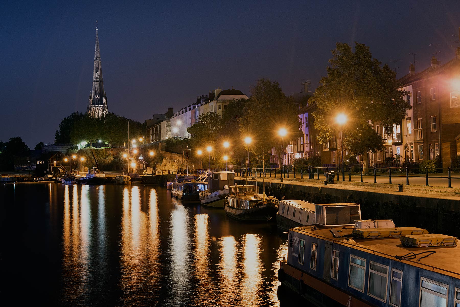 St Mary Redcliffe's spire lit up at night above Bristol's floating harbour, with narrowboats moored along the quay